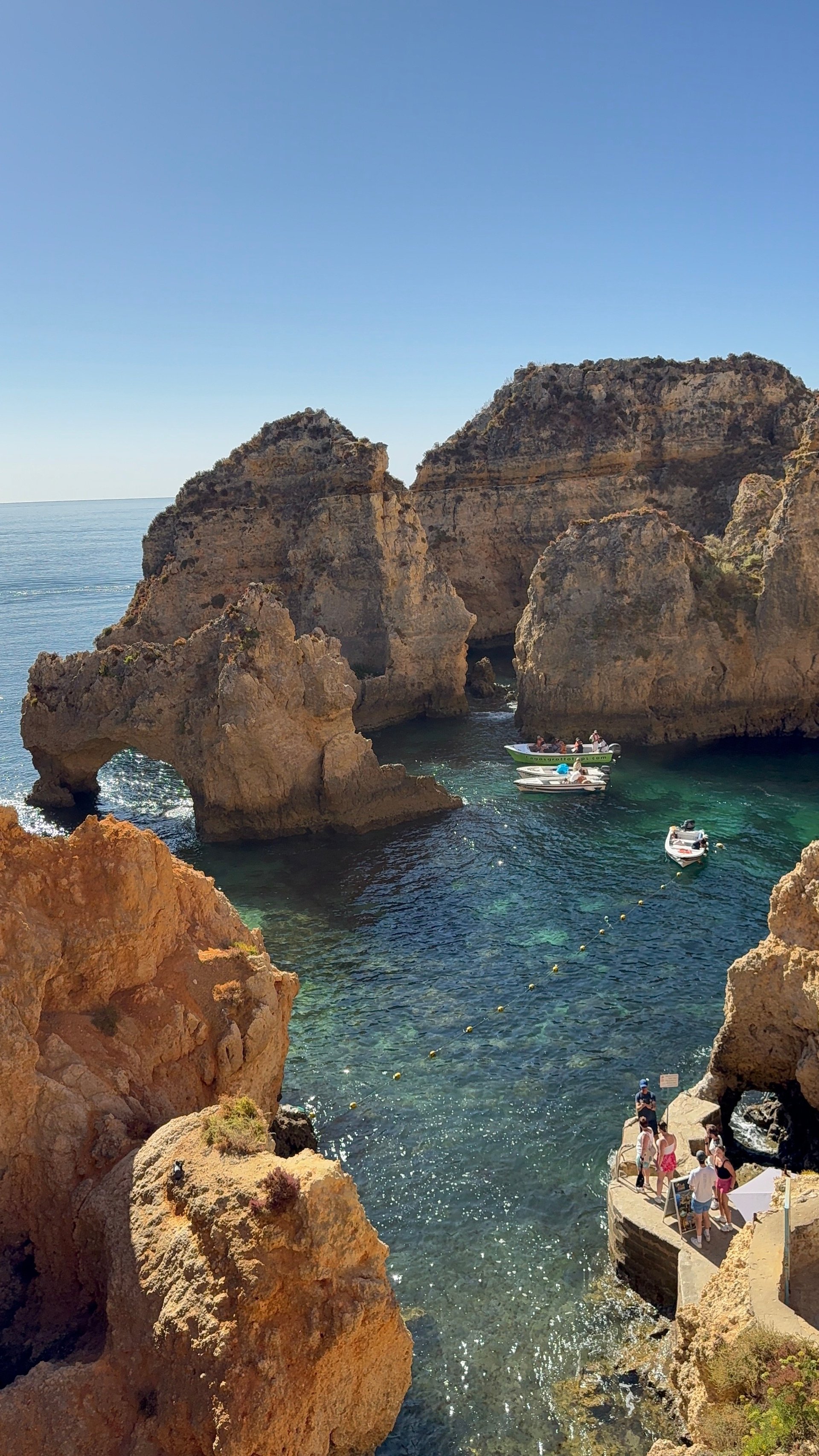 Golden sandstone rock formations and cliffs surrounding a turquoise cove with moored boats and tourists on the ledge