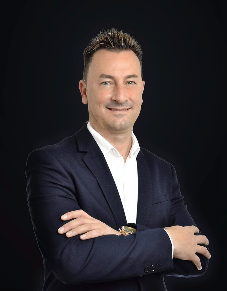 Professional headshot of a man in a dark blazer and white shirt with arms crossed, smiling against a black background